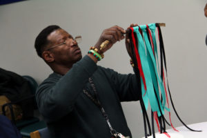 A man examines the colourful fiber art wall hanging he is in the process of creating. The wall hanging consists of ribbons and threads of different lengths and colours, tied to a thin branch.