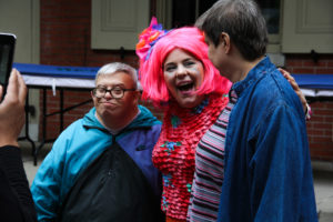 A clown, wearing a bright pink wig and a popcorn shirt, posing for a picture with two Art-Reach members at Art-Reach's 30th birthday party.