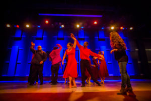 Mixed ability dancers performing on stage. In the center of the dancers is a woman in a red dress with her arm up getting twirled by a man in a matching red shirt