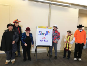 In Photo: Members in Halloween costumes pose with a large notepad that reads "StoryUP! with Art-Reach" Halloween costumes include, a person with a cape and top hat, sparkling jacket with red bow cap, sparkling zebra print jacket with a white safari hat, a hot dog, and a cowboy in a black and orange outfit.