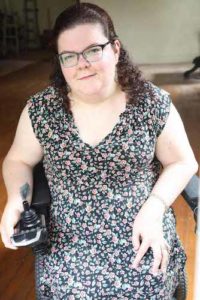 Emily Ladau is a white woman who is wearing glasses and has curly brown hair pulled back halfway, with the rest framing her face. She is wearing a black dress dotted with red flowers and green leaves. She is sitting in a power wheelchair, facing the camera, and smiling. One of her hands is resting in her lap and the other hand is resting on the joystick of her wheelchair, turned so that a tattoo of a peacock feather is visible on her inner arm.