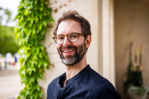 A photo of Tony Derrico, a middle-aged white man with short light brown hair, a full beard and mustache, and tortoiseshell-framed glasses. Tony wears a black button-down shirt and stands in front of a garden wall, smiling.